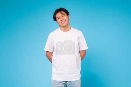 Ein entspannter junger asiatischer Typ in lässiger Kleidung steht mit einem leichten warmen Lächeln vor blauem Hintergrund