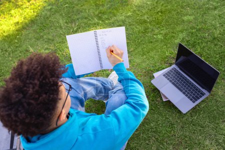 Téléchargez les photos : Vue du dessus de l'étudiant brésilien gars revoir ses notes écrites avec un ordinateur portable ouvert devant lui tout en étant assis sur l'herbe - en image libre de droit