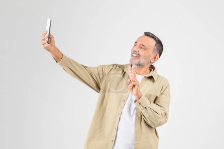 Photo for An older man with grey hair and a beard smiles as he takes a selfie with a white smartphone, dressed in casual attire - Royalty Free Image