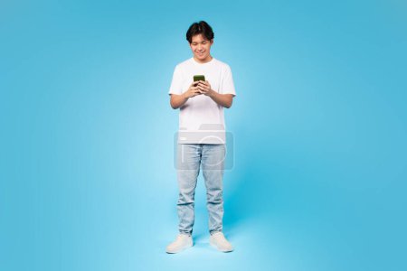 Junge Teenager asiatische Kerl tief in der Verwendung seines Smartphones vertieft, zeigt Technologie Engagement, blauer Hintergrund