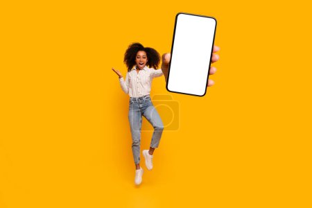 African American woman mid-jump looking excited, holding a smartphone with a blank screen on a vivid yellow background