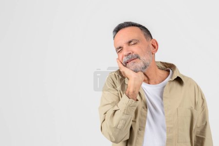A senior man holds his cheek, expressing discomfort, possibly due to a dental issue or toothache, against a clean white background