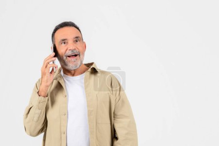 Téléchargez les photos : Un vieil homme joyeux à la barbe grise entame une conversation sur son téléphone portable, exprimant bonheur et surprise - en image libre de droit