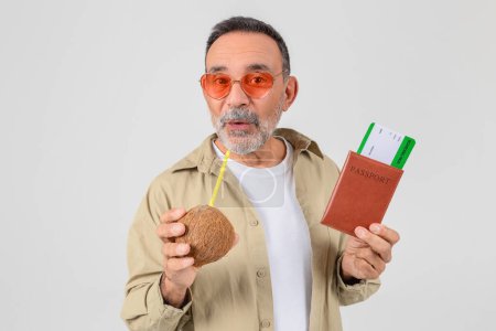 Téléchargez les photos : Joyeux homme âgé en lunettes de soleil funky sirote d'une noix de coco tout en tenant un passeport et une carte d'embarquement, suggérant des vacances tropicales - en image libre de droit