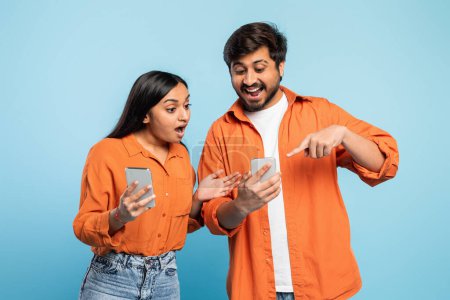 Foto de Sonriente pareja india joven compartiendo y señalando con entusiasmo sus teléfonos inteligentes con un fondo azul - Imagen libre de derechos