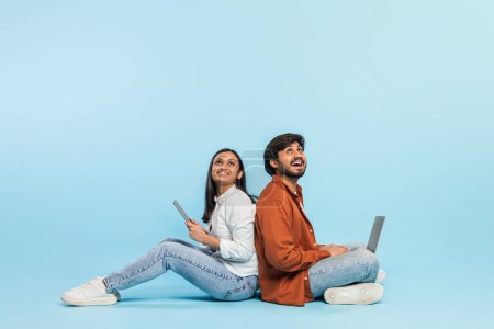 Téléchargez les photos : Assis dos à dos, couple indien s'engage avec la technologie séparément, un homme avec un ordinateur portable et une femme avec une tablette sur un fond bleu - en image libre de droit