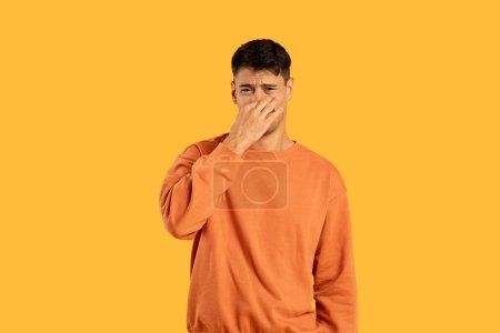 Young man in orange sweater pinching his nose and showing a grimace of disgust on a yellow studio background