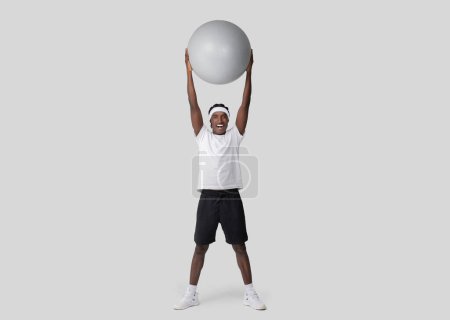 Happy and fit young African American man raises a large exercise ball above the head with excitement on a plain backdrop