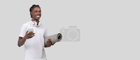 Téléchargez les photos : Un jeune homme afro-américain joyeux tenant un tapis de yoga et un smartphone avec un casque autour du cou sur un fond gris, espace de copie - en image libre de droit