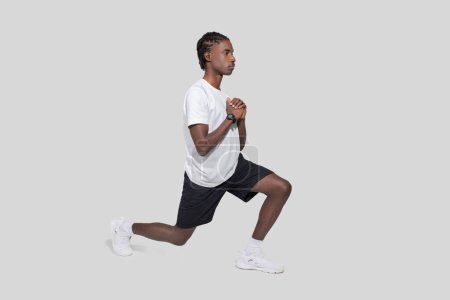 Serious young African American man performs a lunge in workout attire demonstrating exercise form against a gray backdrop