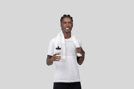 A fit African American man in a white shirt with a towel over his shoulder holds a sports bottle on white background
