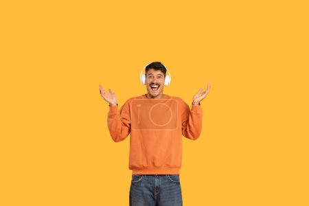 Excited young man with headphones on his head expressing joy and surprise on an orange background, copy space