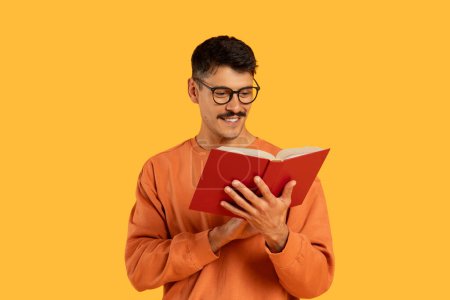 Man in casual clothing with glasses deeply engrossed in reading a red book on orange studio background