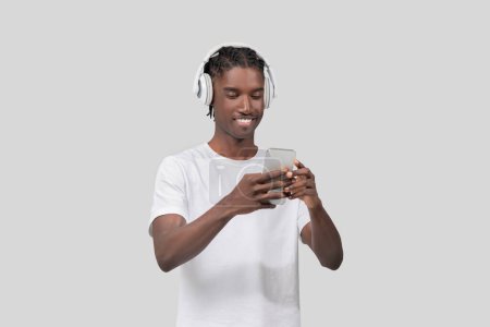 Homme afro-américain décontracté avec écouteurs utilisant un smartphone, profitant de la musique ou podcast sur fond blanc