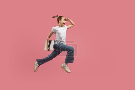 Photo for A young child appears mid-leap with a pencil atop and books underarm against a vibrant pink background, evoking concepts of fun learning - Royalty Free Image