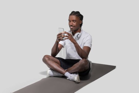 A young athletic African American man in a casual outfit sits on a yoga mat and texts on his smartphone in a studio setting
