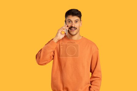 Photo for A cheerful young adult guy in an orange sweatshirt engaging in a pleasant conversation on a mobile phone against a yellow background - Royalty Free Image