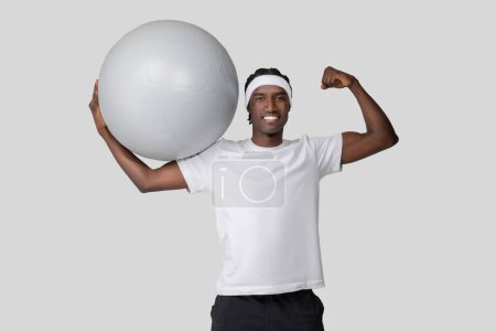 Téléchargez les photos : Un jeune Afro-Américain en forme dans un t-shirt blanc tenant avec confiance une grosse balle d'exercice sur son épaule - en image libre de droit