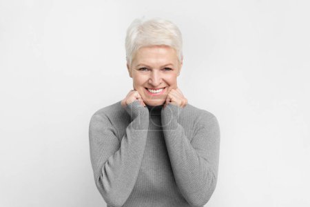 Téléchargez les photos : Joyeuse femme âgée européenne avec un geste ludique frottant ses joues sur fond gris, idéal pour s3niorlife bonheur - en image libre de droit