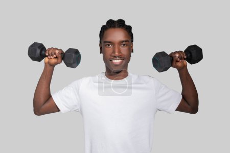 A fit and happy young African American man stands lifting dumbbells while smiling confidently against a neutral backdrop