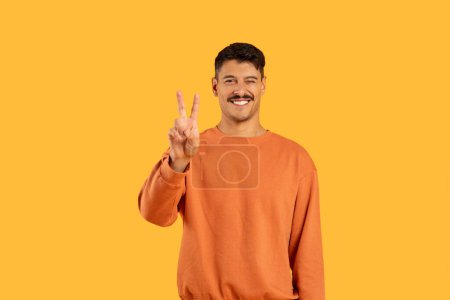 Lächelnder Mann mit Schnurrbart in Freizeitkleidung mit V für Sieg oder Friedenszeichen mit Fingern auf Gelb