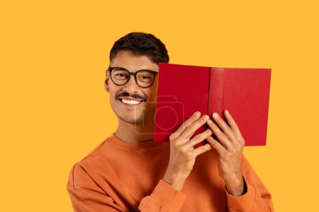 Foto de Hombre alegre con bigote escondido detrás de un libro con una cara sonriente visible sobre un fondo amarillo - Imagen libre de derechos