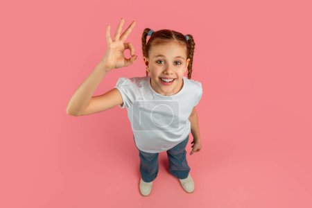 Foto de Jovencita alegre haciendo buen gesto con una mano mirando a la cámara sobre un fondo rosa pastel - Imagen libre de derechos