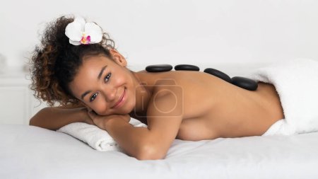 Téléchargez les photos : Femme afro-américaine bénéficiant d'un massage aux pierres chaudes dans un salon de spa - en image libre de droit
