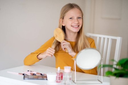 Jovencita alegre cepillándose el pelo largo, sentada en una mesa con espejo y cosméticos, disfrutando del cuidado de la belleza