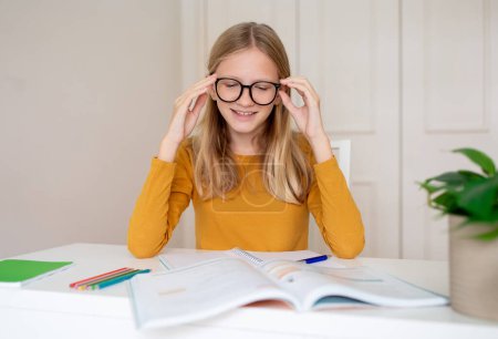 Glückliches Teenager-Mädchen mit Brille, das sich auf sein Studium konzentriert, an einem Schreibtisch mit Büchern und Notizen sitzt, fröhliche Teenager, die es genießen, Hausaufgaben zu machen