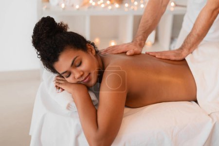 Téléchargez les photos : Contenu jeune femme afro-américaine aux yeux fermés recevant un massage du dos apaisant des mains habiles - en image libre de droit