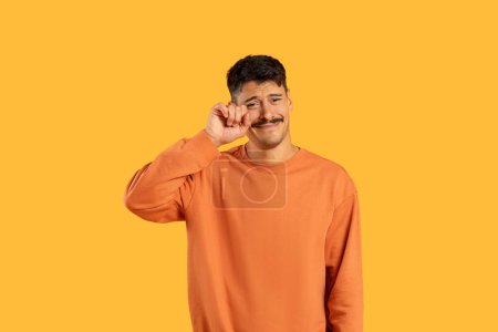 Tearful emotional man with moustache in an orange sweater wiping away a tear on a yellow background