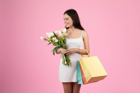 Téléchargez les photos : Une jeune femme joyeuse au look européen tient des fleurs printanières et des sacs à provisions, évoquant une ambiance estivale, parfaite pour le contenu isolé de la génération Z - en image libre de droit
