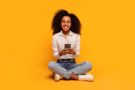 Téléchargez les photos : Contenu jeune femme afro-américaine assise avec les jambes croisées à l'aide d'un smartphone souriant à la caméra, fond jaune - en image libre de droit