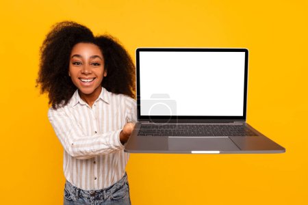 Téléchargez les photos : Souriante jeune femme afro-américaine présentant un ordinateur portable ouvert moderne avec une maquette d'écran blanc vierge - en image libre de droit