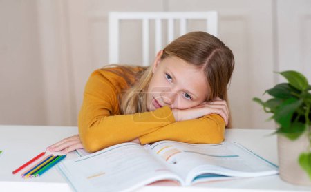 Bored young girl sitting at a table with book, feeling tired after doing homework, upset female teenager resting head on hands and looking at camera