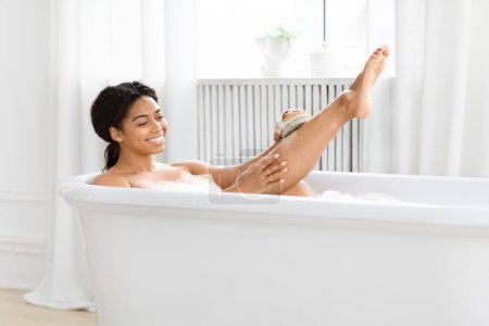 Foto de Juguetona joven afroamericana fregando sus piernas con cepillo mientras disfruta de un baño de burbujas en casa - Imagen libre de derechos