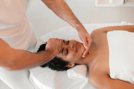 Téléchargez les photos : Un masseur professionnel offrant un massage relaxant du visage à la dame afro-américaine avec serviette couvrant le bas du corps dans un cadre de spa serein - en image libre de droit