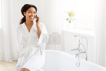 Foto de Mujer afroamericana sonriente disfruta de una llamada telefónica con una bata blanca posada en el borde de una bañera en una habitación luminosa - Imagen libre de derechos