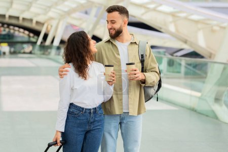 Téléchargez les photos : Couple heureux marchant dans un aéroport avec café à emporter, homme et femme joyeux portant des bagages et se regardant, appréciant voyager ensemble - en image libre de droit