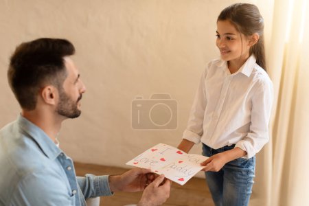 Téléchargez les photos : Une jeune fille remet une carte I Love You Daddy à son père, montrant de l'affection dans une pièce éclairée - en image libre de droit