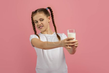 Chica joven repugnante mira un vaso de leche que sostiene, sobre un fondo rosado, no le gustan los productos lácteos