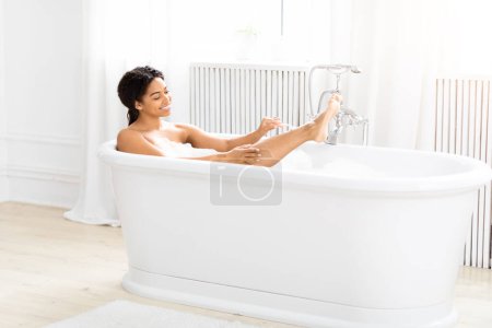 A joyful African American young woman enjoys a relaxing bubble bath in a white, freestanding bathtub in a bright, clean bathroom