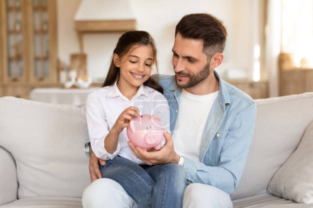 Father teaching daughter about savings, both smiling as they insert money into a piggy bank while seated