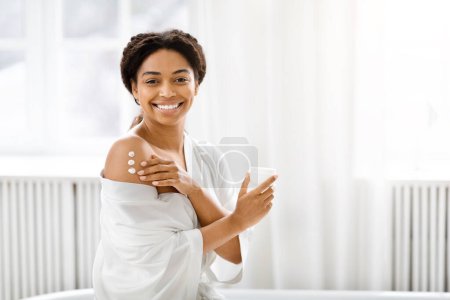 Téléchargez les photos : Femme afro-américaine souriante en peignoir blanc tient un produit de soins de la peau, incarnant l'autosoin et la routine de beauté quotidienne - en image libre de droit