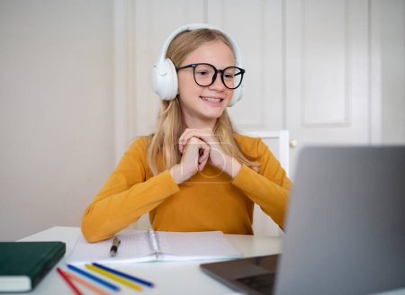 Téléchargez les photos : Une jeune étudiante heureuse avec des lunettes et des écouteurs assis à un bureau, participant à une classe virtuelle, une adolescente étudie à la maison - en image libre de droit