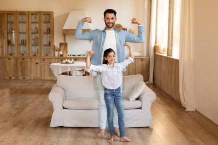 Foto de Padre e hija joven flexionando sus músculos juntos, compartiendo un momento de fuerza y diversión en casa - Imagen libre de derechos