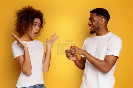 Photo for Surprising proposal. Shocked african american woman looking at her boyfriend with engagement ring, orange background - Royalty Free Image