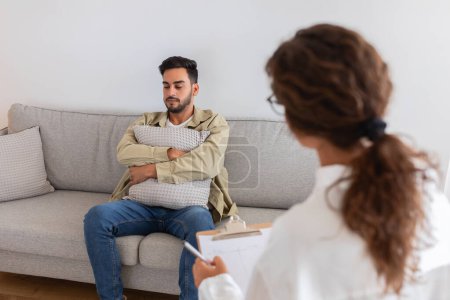 In a therapy session, middle eastern man seeking help clutches a pillow while talking to psychologist, conveying vulnerability