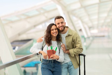Foto de Un hombre y una mujer sonrientes de pie en una moderna terminal del aeropuerto, con pasaportes y billetes, jóvenes cónyuges felices disfrutando viajando juntos, espacio para copiar - Imagen libre de derechos